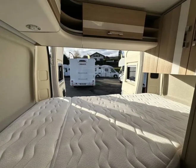 Intérieur d'un camping-car avec lit et placards en bois, vue sur d'autres véhicules stationnés.