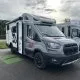 Camping-car gris stationné sur un parking, porte ouverte, ciel nuageux en arrière-plan.