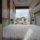 Intérieur d'un camping-car avec lit et rangements, porte arrière ouverte sur un parking.