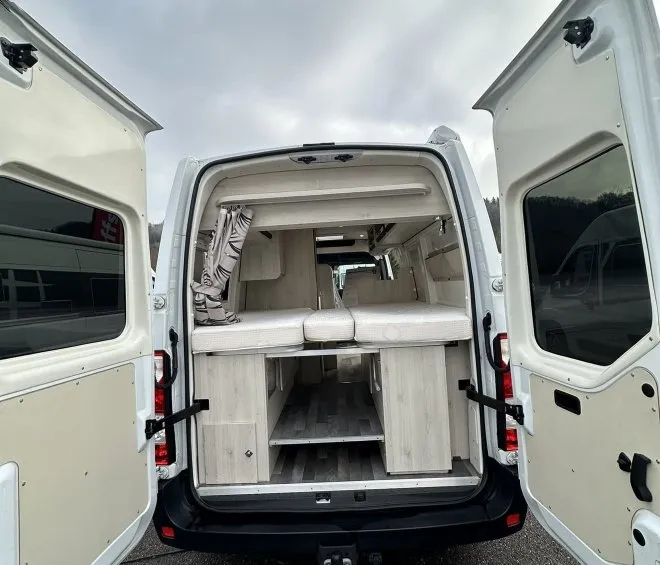 Intérieur aménagé d'un van avec portes arrière ouvertes, montrant des rangements et un espace de couchage.