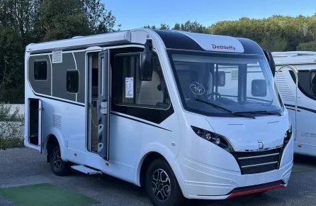 Camping-car Dethleffs blanc stationné sur un parking, porte ouverte, ciel bleu en arrière-plan.
