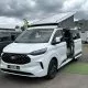 Van aménagé Ford blanc avec toit relevable sur un parking de concessionnaire.