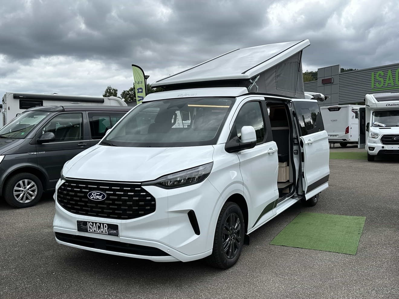 Van aménagé Ford blanc avec toit relevable sur un parking de concessionnaire.