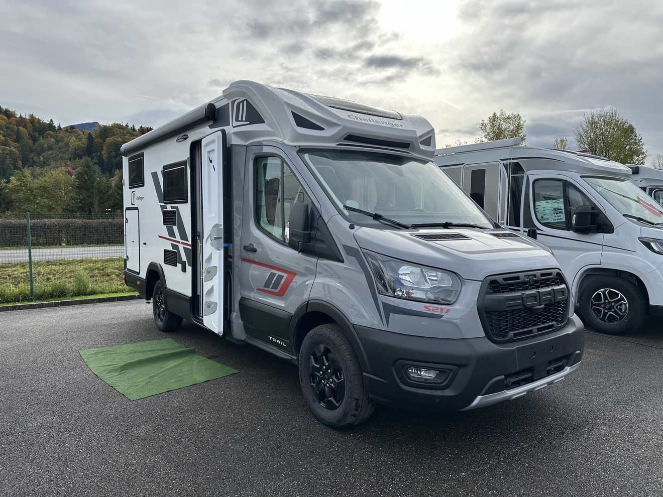 Camping-car gris stationné sur un parking, porte ouverte, ciel nuageux en arrière-plan.