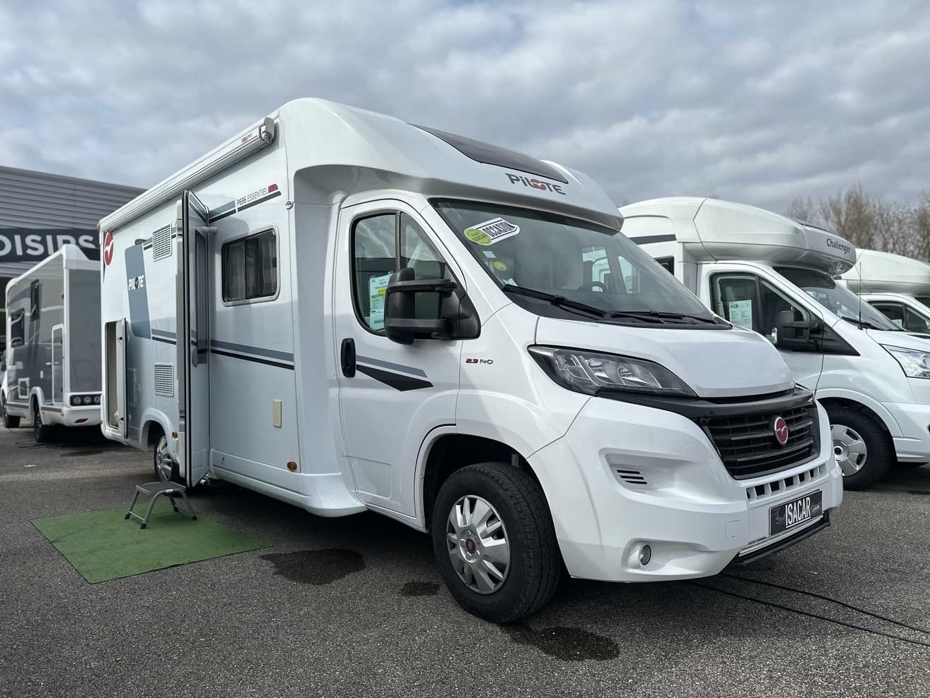 Camping-car blanc stationné sur un parking, modèle Pilote, ciel nuageux en arrière-plan.